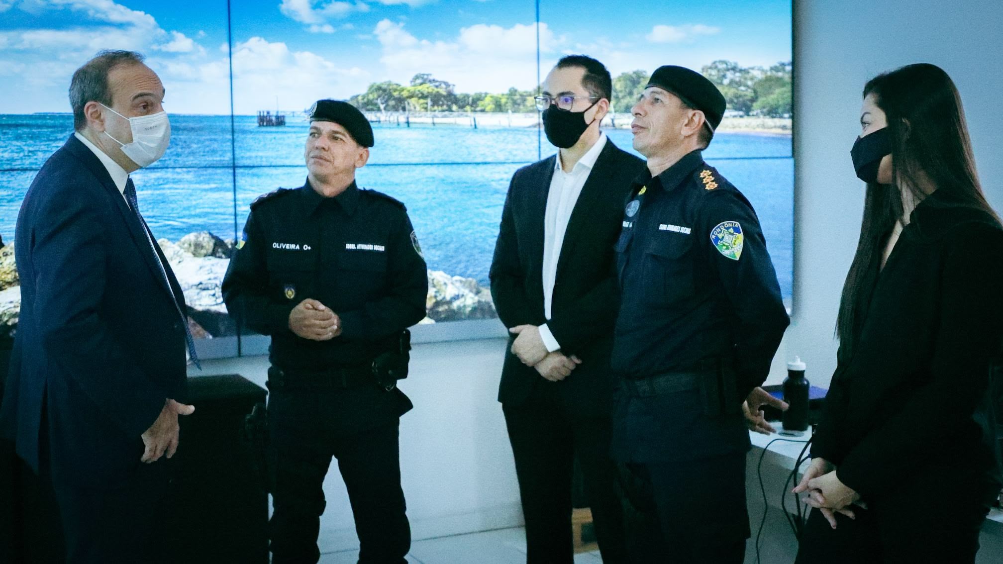 A fotografia mostra um homem de terno falando com 4 pessoas, duas delas estão vestidas com uniformes de policiais