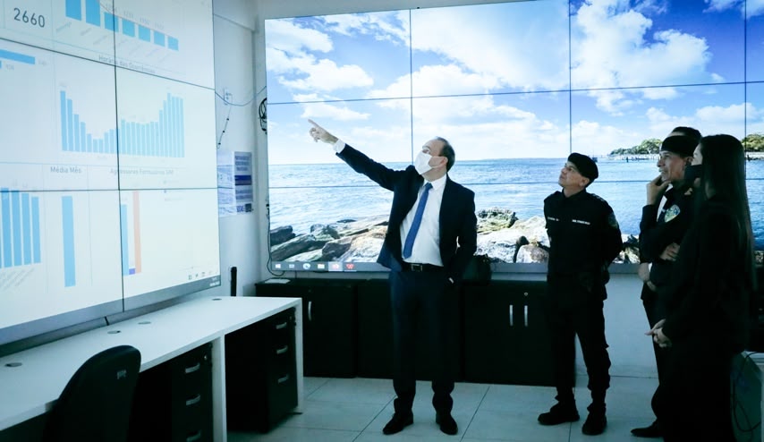 A fotografia mostra um homem de terno apontando para um telão, e atrás dele estão 4 pessoas vestidas com uniformes de policiais